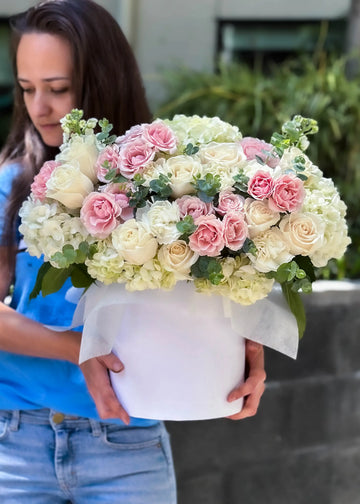 Rose and hydrangea arrangement of white roses and pink spray roses in a white hat box, held in both hands