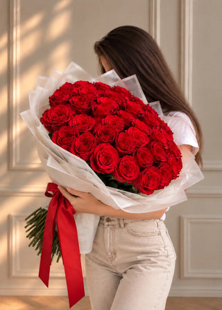 Red roses bouquet in layered paper with a long satin ribbon, held in both arms against a paneled cream wall