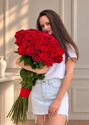 Long stem red roses held in both arms, bare stems bound with a ribbon, against a paneled cream wall
