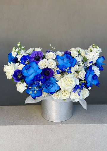 Blue and white flower arrangement of blue roses, white roses, purple anemones and delphinium in a silver box