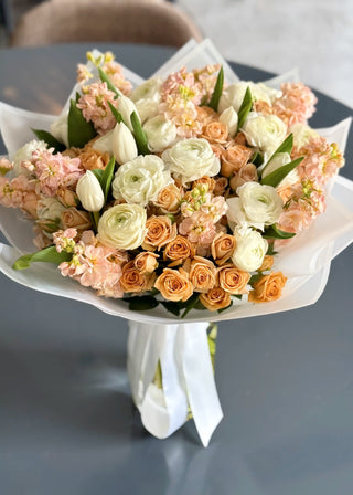 Pastel flower bouquet with peach spray roses, white ranunculus, ivory tulips and pink stock blooms, wrapped in white paper and ribbon.