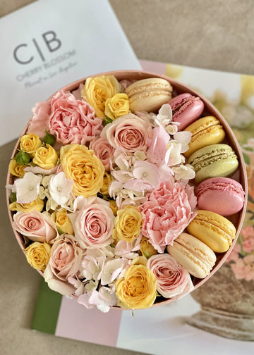 Round pink box of flowers and macarons: blush and yellow spray roses, carnations and hydrangea, top view