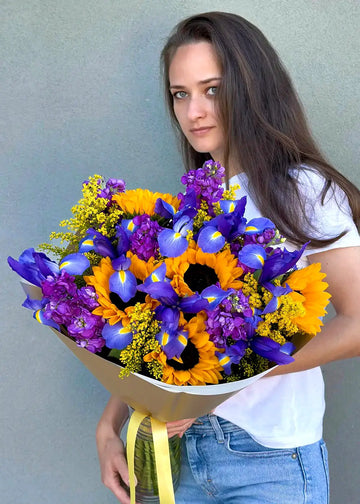 Bouquet of sunflowers with purple flowers, blue irises, purple stock and solidago in paper wrap, held in arms
