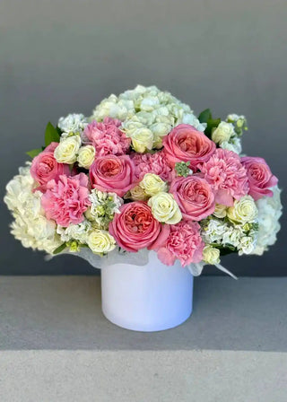 White round flower box overflowing with pink peony roses, ivory spray roses, white hydrangeas and pale pink carnations in a lush arrangement.