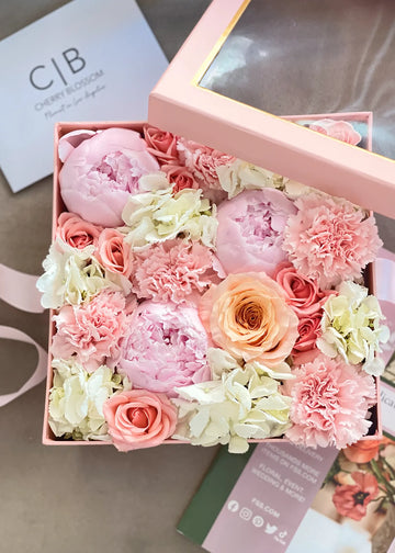 Square box flower arrangement of pink peonies, carnations, coral spray roses and cream hydrangea, lid propped open