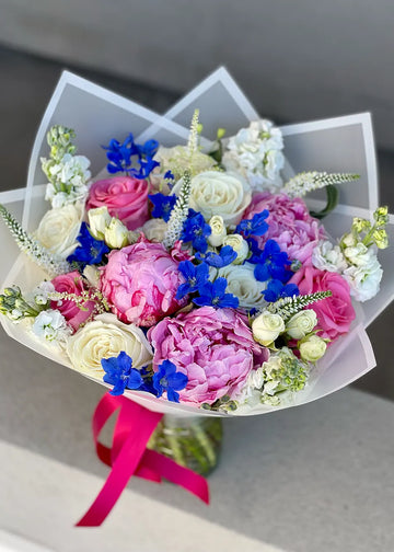 Peony and rose bouquet with pink peonies, white roses, blue delphinium and white stock in paper wrap with a ribbon