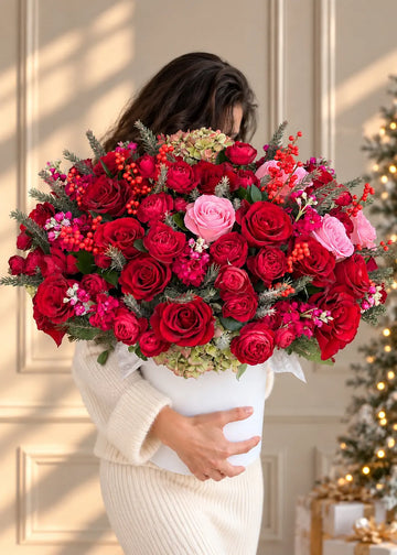 Woman holding a large Christmas flower box of red roses, pink roses and ilex berries beside a lit Christmas tree