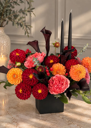 Close view of crimson and orange pompon dahlias, coral garden roses and red berries around two black taper candles