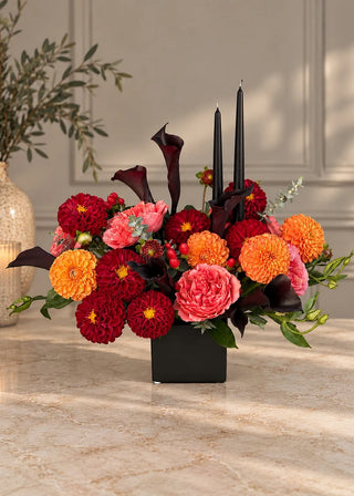 Fall centerpiece of red and orange dahlias, coral garden roses and black calla lilies with two black candles, front view