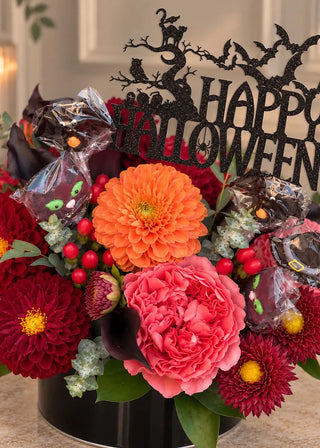 Close view of an orange dahlia, coral garden rose and red berries with cat and bat lollipops under a Happy Halloween topper