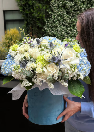 Blue hydrangea arrangement with white roses, sea holly and green carnations in a blue box, held in hands