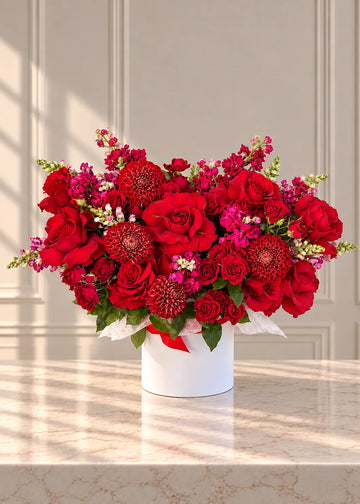 Round white box of red roses, red pompon dahlias, magenta stock and snapdragons on a marble counter