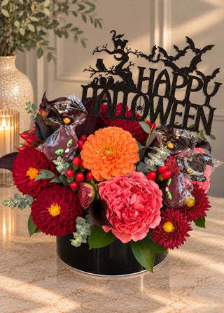 Halloween flowers with candy: red and orange dahlias, a coral garden rose and lollipops in a round black tin, front view