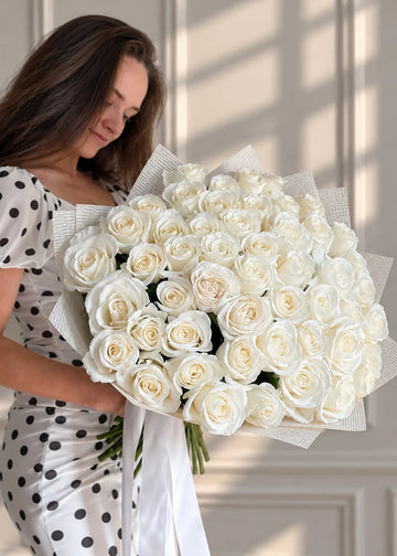 White roses bouquet wrapped in paper with a long ribbon, held in a woman's arms against a paneled wall