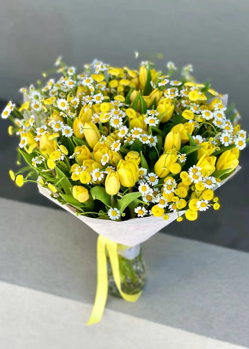 Round bouquet of 40 yellow tulips with white chamomile and yellow button chamomile in a glass vase on a concrete ledge